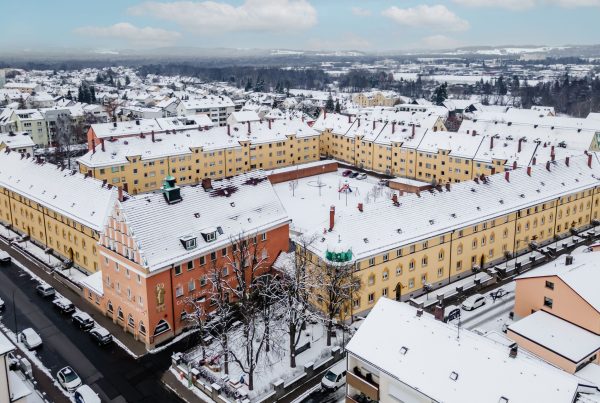 sehr großer Wohnblock aus der Vogelperspektive. Ein Gebäude ist orange die restlichen gelb. Innenhof ist zu sehen mit Spielplatz und Stellplätze. Es liegt Schnee.