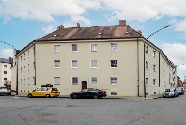 Wohnblock, drei Reihen an Fenster mit weißer Umrandung zu sehen. Direkt an der Straße mit parkenden Autos davor. Eingangstür genau in der Mitte.