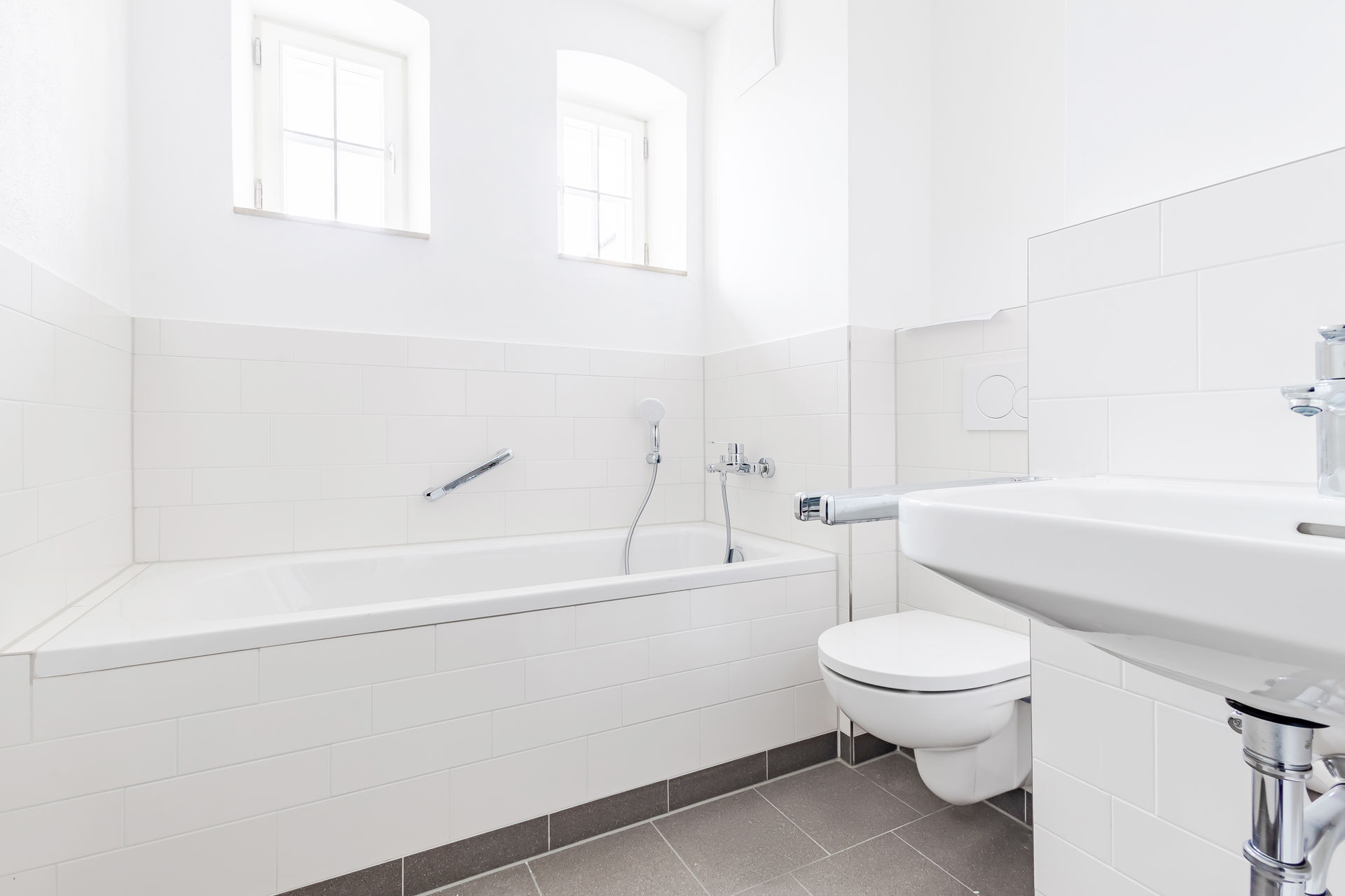 helles Badezimmer, zwei kleine Fenster ganz oben im Raum, gleich darunter ine große Badewanne mit Toilette gleich davor und Waschbecken.