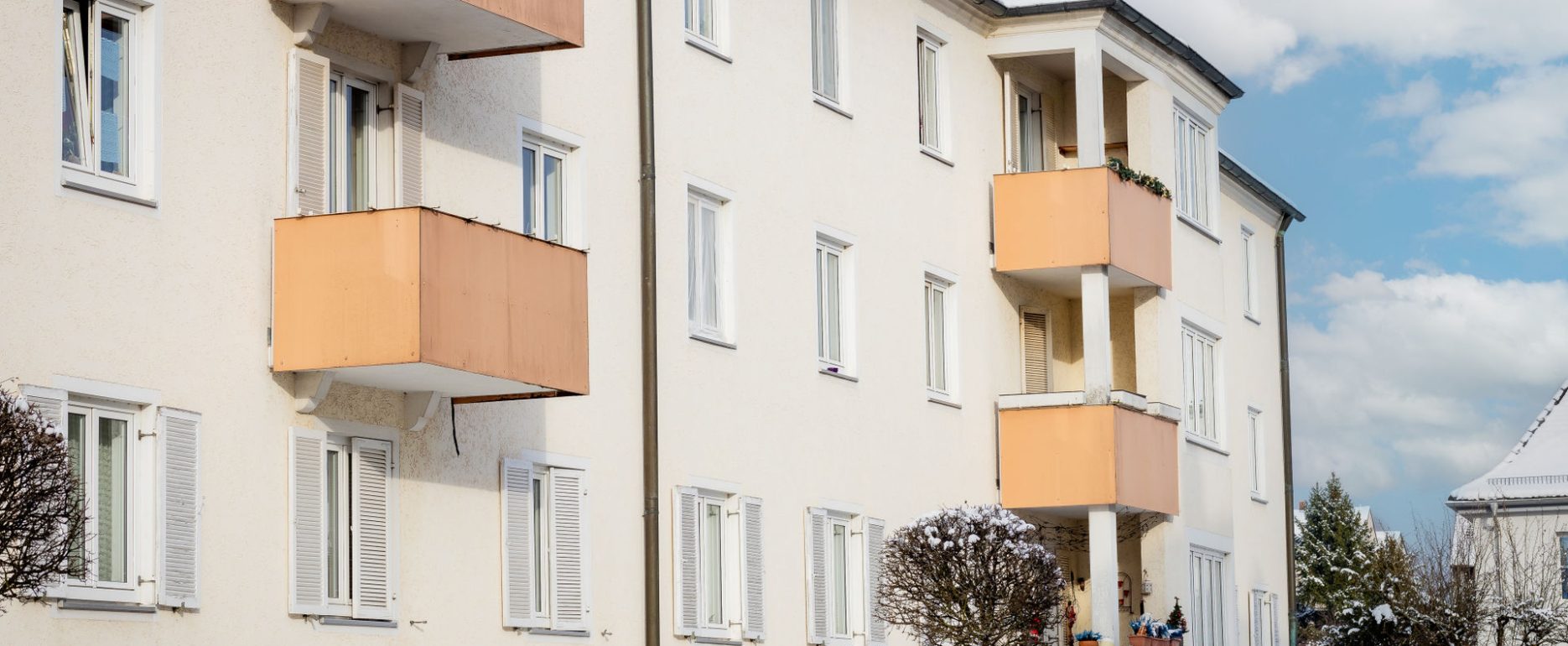 Ansicht Mehrfamilienhaus, 3 Stockwerke mit 5 Balkone in orange. Fassade ist hell und die untersten Fenster haben weiße Holzläden. Vor dem Wohnblock befindet sich ein Gartenzaun mit Bepflanzung. Es ist Winter.