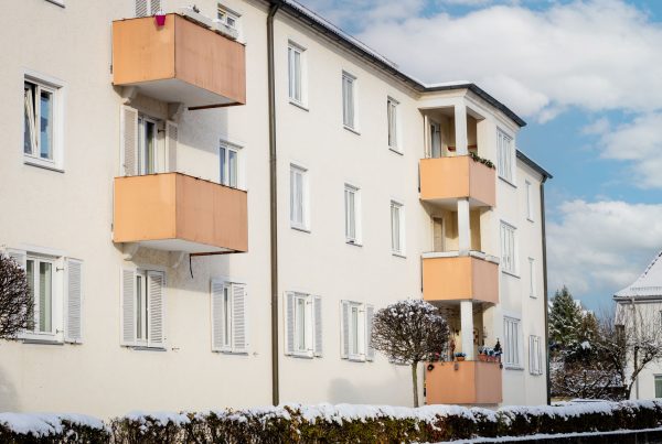 Ansicht Mehrfamilienhaus, 3 Stockwerke mit 5 Balkone in orange. Fassade ist hell und die untersten Fenster haben weiße Holzläden. Vor dem Wohnblock befindet sich ein Gartenzaun mit Bepflanzung. Es ist Winter.